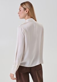Blusa di seta color crema con colletto alto e maniche lunghe, dotata di polsini con bottone. Texture morbida e vestibilità rilassata, abbinata a pantaloni marroni.