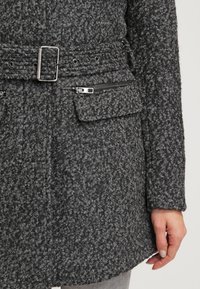 Gros plan d'un manteau en laine gris foncé texturé avec ceinture, boucle métallique, poche zippée, poche à rabat et une main reposant sur le côté.