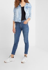 Ljusblå denimjacka, svart åtsittande topp och högmidjade blå jeans med smal passform, kombinerat med vita sneakers med gröna accenter.