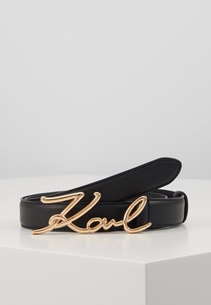 Czarny skórzany pasek z błyszczącą złotą klamrą z napisem "Karl" kursywą, zaprezentowany zwinięty na białej powierzchni na szarym tle.