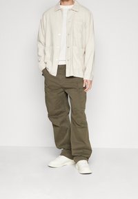 Lichtbeige button-up shirt met zakken, gecombineerd met olijfgroene cargobroek met zijzakken, en witte sneakers. Eenvoudig ontwerp en comfortabele pasvorm.