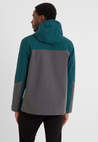 Man wearing a color-block jacket in teal and gray with a hood, shown from the back, against a plain light background.