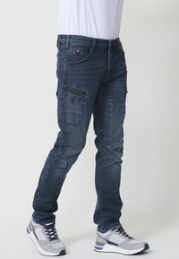 Pantalones cargo de denim en azul oscuro con múltiples bolsillos, tejido texturizado, diseño ajustado y detalle de cierre con cremallera. Combinados con zapatillas deportivas grises.
