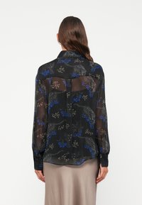 Blusa nera trasparente con stampa floreale e animale colorata. Caratterizzata da maniche lunghe, polsini con bottoni e orlo curvo. Tessuto leggero.