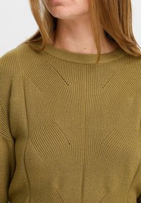 Olíva zöld kötött pulóver, amely textúrált mintázattal rendelkezik, függőleges vonalakkal és V-alakú díszekkel. Bordás nyakú és laza fazonú.