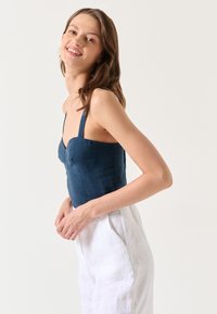 Niebieski lniany crop top z regulowanymi ramiączkami, dopasowanym korpusem i dekoltem w kształcie serca, w zestawie z białymi lnianymi spodniami. Gładka tekstura.