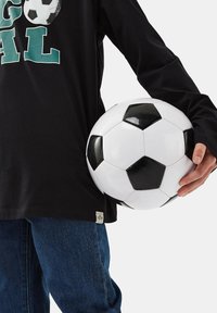 Schwarzes Langarmshirt mit grüner Schrift, getragen mit blauen Jeans; hält einen schwarz-weißen Fußball mit glänzender Oberfläche und sechseckigen Panels.