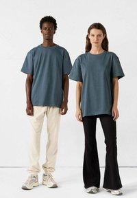 Due persone che indossano t-shirt a taglio rilassato in tessuto blu ardesia, abbinate a pantaloni svasati di colore chiaro e neri, che stanno fianco a fianco.