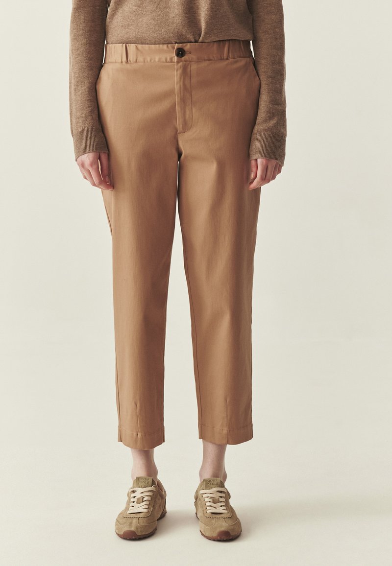 Osoba ubrana w krótkie camelowe spodnie, brązowy top z długim rękawem i beżowe sneakersy z białymi sznurówkami, stojąca na neutralnym tle.