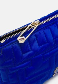 Blå quiltet nylon taske med en gylden lynlås og hardware. Har en mønstret tekstur med en rund detalje nær lynlåsens træk.