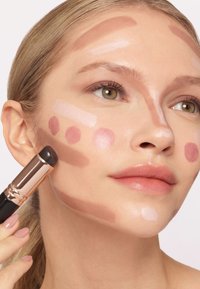 Stick de maquillaje aplicando varias tonalidades de crema en la piel: swatches de rosa claro, marrón oscuro y beige suave en la mejilla; textura suave; acabado luminoso.