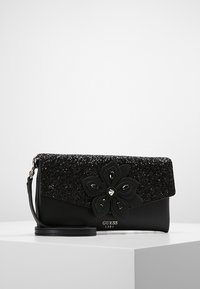 Sac à main noir avec un rabat pailleté noir, orné d'un grand accent floral. Texture lisse, bandoulière détachable et logo de marque métallique.