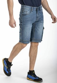 Shorts cargo en denim bleu clair, avec de multiples poches, des coutures double couture et une coupe décontractée. Portés avec des chaussures de sport noires et bleues.