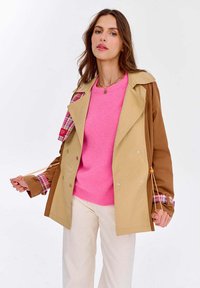 Bežovo-hnedý trench coat s károvanými akcentmi na manžetách, striebornými gombíkmi a s nastaviteľnou šnúrkou. Nosený cez ružový sveter a krémové nohavice.