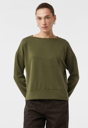 Donna con i capelli raccolti, che indossa orecchini a cerchio argentati, una maglietta a maniche lunghe verde oliva e pantaloni marroni scuri, in piedi davanti a uno sfondo bianco.