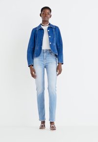 Chaqueta de denim en azul oscuro, con un collar y botones blancos; combinada con jeans ajustados en azul claro y sandalias blancas con tiras.