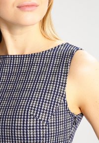 Robe en tissu texturé avec un motif pied-de-poule bleu marine, noir et blanc ; présente un décolleté arrondi et un design sans manches avec des coutures visibles.