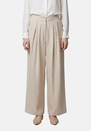 Frau trägt beige, hoch taillierte, weite Hosen mit Plisseefalten, eine weiße langärmelige Bluse und beigefarbene spitze Absatzschuhe.