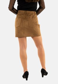 Brauner Wildleder-Minirock mit tailliertem Bund und zwei Gesäßtaschen. Das Modell trägt schwarze High Heels, die eine glatte Textur zur Schau stellen.