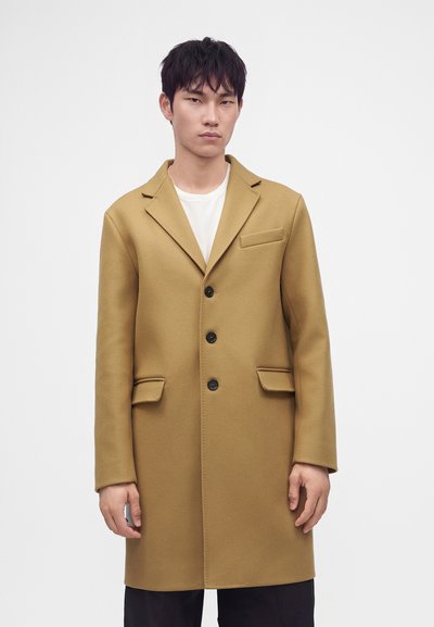 Jeune homme debout portant un manteau camel boutonné par-dessus une chemise blanche sur un fond blanc uni.
