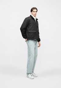 Casaco preto com fecho de correr, com textura macia e dois bolsos frontais, combinado com jeans azul claro e ténis brancos. Modelo em perfil.