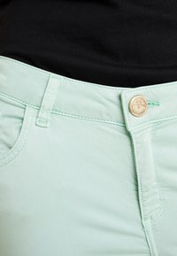 Light green cotton pants with a zip fly, button closure, slanted pockets, and contrasting turquoise stitching. The fabric appears smooth.