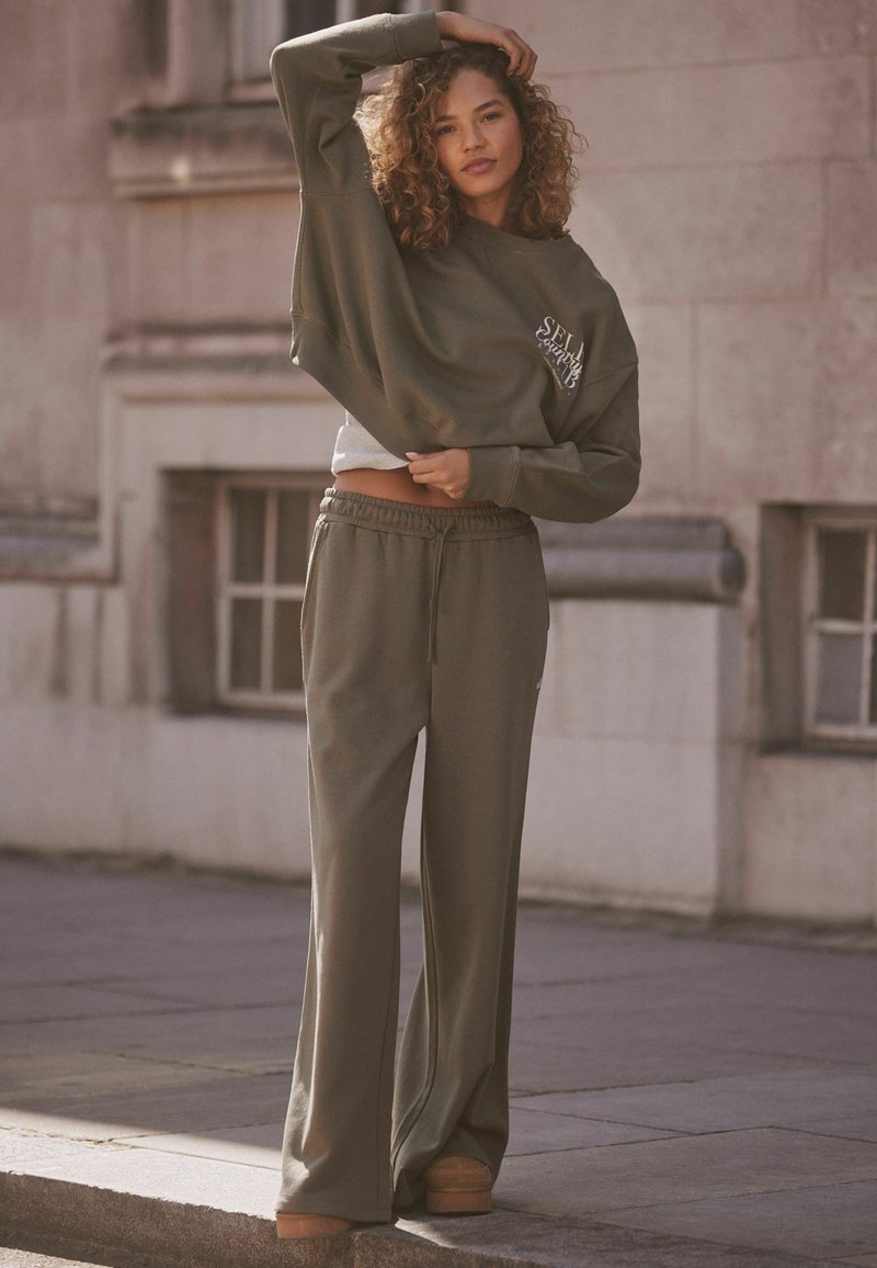 Felpa verde oliva a vita corta con logo, abbinata a pantaloni larghi verde oliva e scarpe con platform beige. Tessuto morbido e vestibilità rilassata.