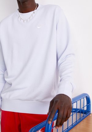 Person trägt weißen Nike-Sweatshirt und rote Hose, legt Hand auf die Rückenlehne eines blauen Metallstuhls vor weißem Hintergrund.