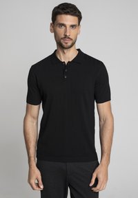 Mężczyzna z krótkimi brązowymi włosami i brodą, ubrany w czarną koszulę polo z krótkim rękawem oraz ciemne spodnie, stojący na szarym tle.