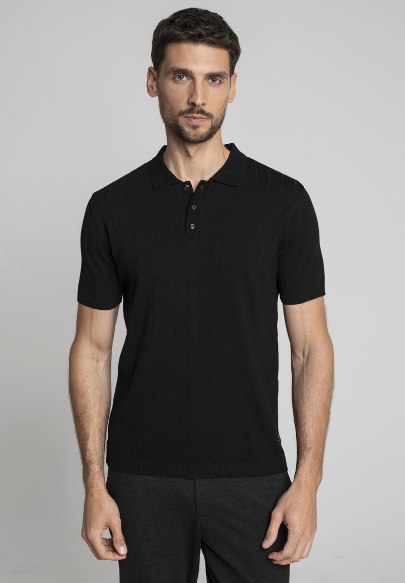 Mężczyzna z krótkimi brązowymi włosami i brodą, ubrany w czarną koszulę polo z krótkim rękawem oraz ciemne spodnie, stojący na szarym tle.