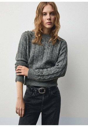 Grauer Pullover mit Kabelstrickmuster, verziert mit kleinen Perlen auf den Schultern. Kombiniert mit hochgekrempelten schwarzen Jeans und einem Gürtel.
