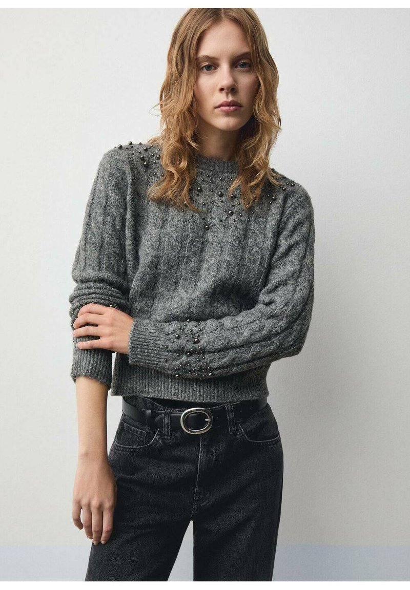 Maglione grigio con texture a treccia, adornato con piccole perline sulle spalle. Abbinato a jeans neri a vita alta e una cintura.