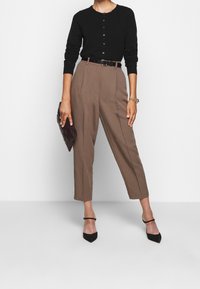Cardigan nero a maniche lunghe, pantaloni marroni a vita alta, cintura nera, tacchi neri con punta affilata; in mano una pochette scura; design minimalista.