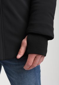 Melna jakna ar gludu tekstūru, kurai ir ribots piedurknes, kur atpūšas roka. Tumši zili džinsi daļēji redzami.