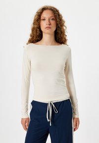 Haut long beige clair à col bateau et design ajusté, associé à un pantalon bleu marine avec cordon de serrage et accents rayés contrastants.
