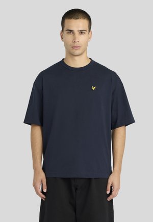 Jeune homme aux cheveux courts portant un T-shirt oversize bleu marine avec un petit logo d’oiseau jaune sur la poitrine gauche.