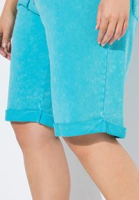 Shorts en denim turquoise présentant une surface douce et texturée ainsi que des ourlets roulés. Mettant en valeur un design épuré et des détails de couture visibles.