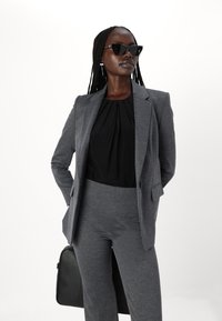 Gray tailored suit with a fitted blazer and high-waisted trousers, paired with a black blouse. Accessorized with black sunglasses and a black bag.