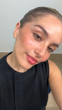 Nahaufnahme einer Person mit heller Haut und braunen Augen, die ein schwarzes ärmelloses Oberteil trägt, mit natürlichem Make-up, das glänzende Lippen und einen subtilen Rouge-Ton beinhaltet.
