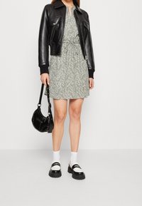 Femme portant une robe florale verte avec une taille nouée, une veste en cuir noire, des chaussettes blanches, des mocassins noir et blanc, et tenant un sac à main noir.