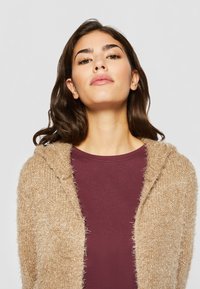 Hettejakke i beige, fluffy materiale med teksturert design. Under er en dyp burgunder tettsittende topp synlig.