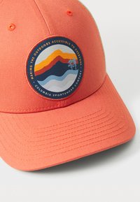 Coral baseball cap with a round logo featuring layered mountains in orange, blue, and white. Text around the logo highlights accessibility.