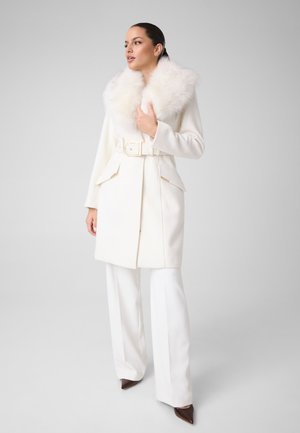 Femme portant un manteau blanc ceinturé avec un grand col duveteux, un pantalon blanc à jambes larges et des chaussures marron pointues, debout devant un fond gris.
