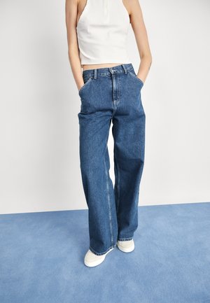 Personne portant un jean bleu taille haute à jambes larges, un crop top blanc sans manches et des chaussures blanches, debout sur un tapis bleu, les mains dans les poches.