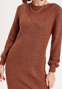 Robe tricotée marron à manches longues, présentant une texture côtelée et un décolleté en V. Inclut un collier en chaîne en accent.