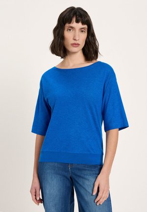 Frau mit schulterlangem, dunklem Haar, die ein hellblaues Halbarm-Oberteil und mittelblaue Jeans trägt, stehend vor einem einfarbigen hellen Hintergrund.