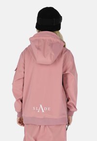 Roze jasje met een capuchon, gemaakt van gladde stof. Beschikt over geribbelde manchetten en een logo op de achterkant. Gecombineerd met een zwarte beanie.