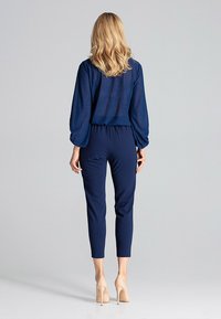 Granatowy kombinezon z dopasowanym talią i zwężanymi nogawkami, z przezroczystymi długimi rękawami oraz fakturowanym detalem materiału. Połączony z cielistymi szpilkami.