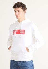 Hanorac alb dintr-un material moale, cu buzunar frontal și glugă cu șnur. Design de logo dreptunghiular roșu și albastru pe piept.
