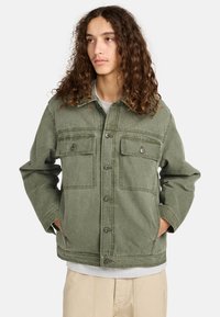 Chaqueta de mezclilla verde oliva con dos bolsillos en el pecho, cierre de botón en el frente y un corte relajado. Cuello redondeado y mangas largas con textura mínima.